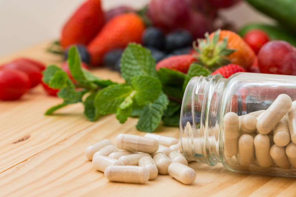  Cápsulas de suplemento com frutas ao fundo, ilustrando fontes naturais e sintéticas de vitaminas hidrossolúveis e sua importância para a saúde.