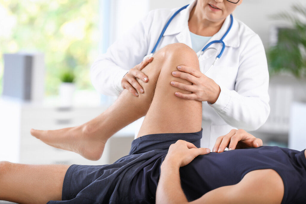 Pessoa deitada com perna direita levantada e dobrada, enquanto médica segura o joelho do paciente com ambas as mãos, indicando dor no joelho ao dobrar
